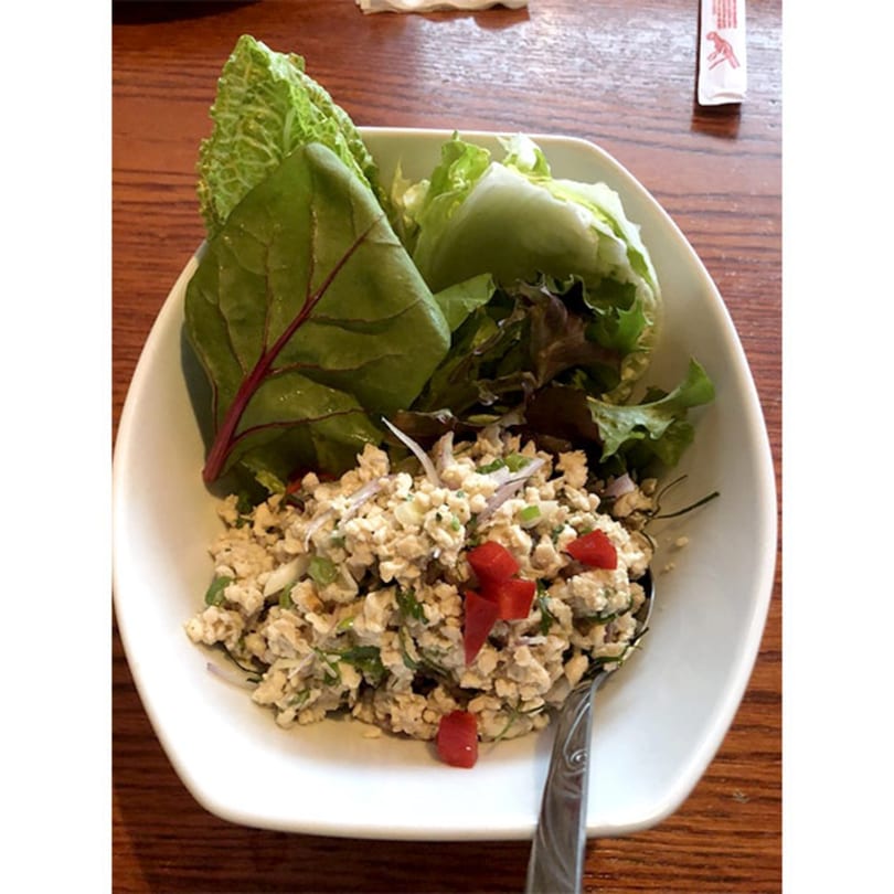 A white bowl of Thai larb with minced meat, red peppers, and lettuce wraps on a wooden table.
