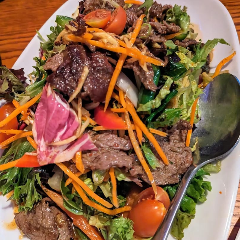 Tong Thai beef salad with mixed greens, carrots, tomatoes, and a spoon on a white plate.