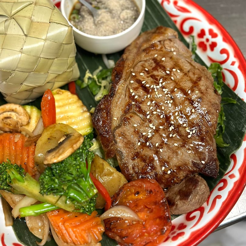 Grilled steak with sesame seeds, mixed vegetables, and a woven basket of sticky rice on a Thai plate.