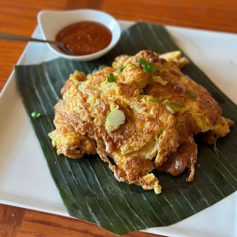 Fluffy golden Thai omelets with green onions on a banana leaf, served with chili sauce.