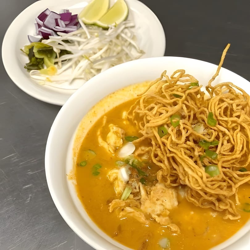 A bowl of Tong Thai's Khao Soi with crispy noodles, chicken, and a side of fresh garnishes.